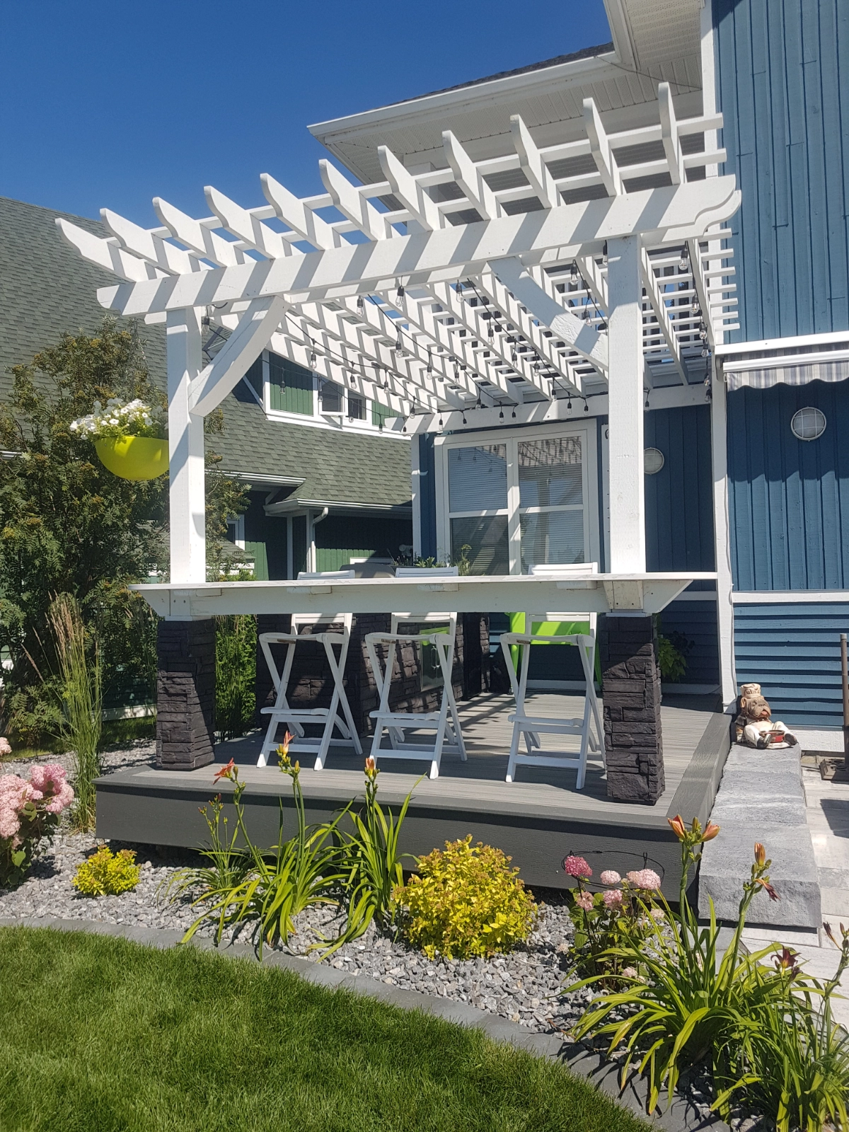 white wood pergola with outdoor kitchen and patio bar, surrounded by beds with flowers and shrubs