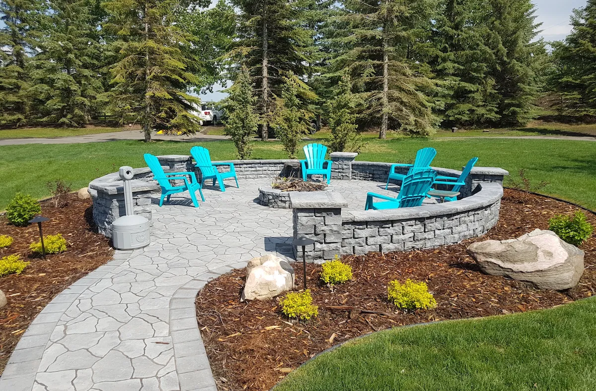 Concrete path to round patio with firepit and pillar wall surrounded by landscaping of large rocks and bushes
