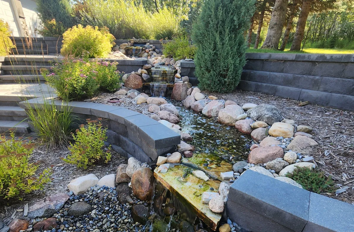 Tiered Keystone retaining walls with water feature running through. Surrounded by flourishing shrubs and grasses.