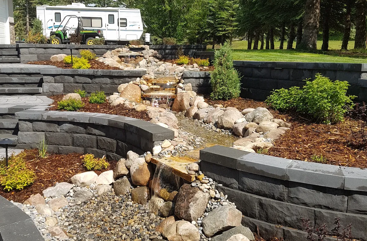 Tiered Keystone retaining walls with water feature running through. Surrounded by newly planted shrubs.